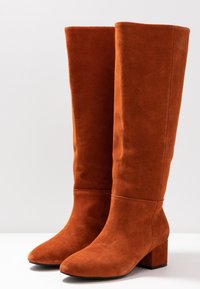 Botas de ante naranjas hasta la rodilla con tacón cuadrado, punta afilada y textura suave. Diseño minimalista con una línea clara en la parte superior.