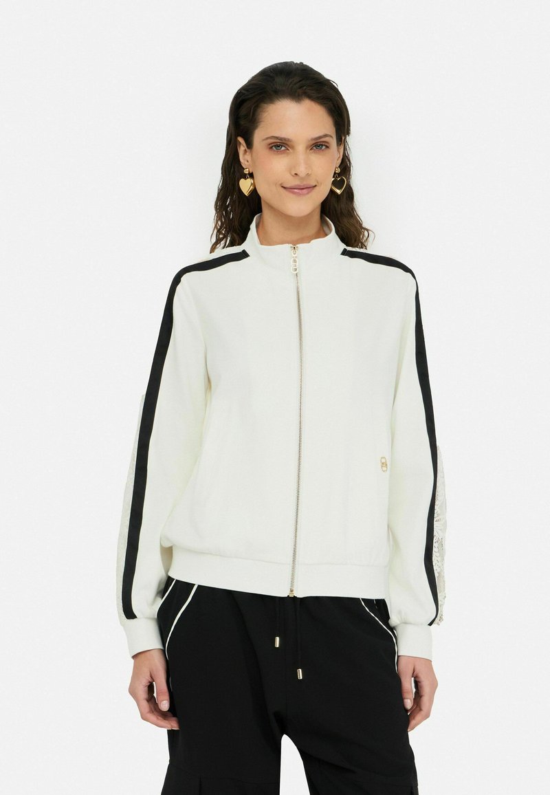 Veste blanche à zip avec des rayures noires sur les côtés et des détails en dentelle sur les manches, dotée d'un col montant et de poignets côtelés. Conçue pour un usage décontracté.
