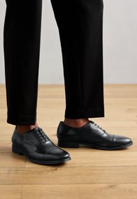 Zapatos de vestir de cuero negro con punta redondeada, diseño con cordones y detalles de costura sutiles, combinados con pantalones negros a medida.