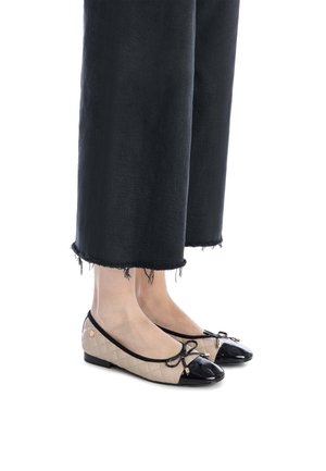 Zapatos planos acolchados beige con punteras de charol negro y acentos de lazo, combinados con pantalones de pierna ancha negros deshilachados.