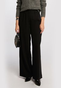 Black wide-leg trousers with an elastic waistband, made of smooth fabric, paired with a textured gray cardigan and a small black handbag.