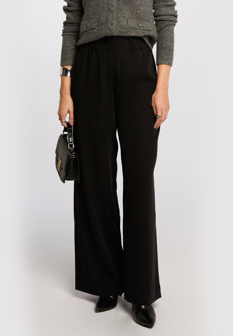 Black wide-leg trousers with an elastic waistband, made of smooth fabric, paired with a textured gray cardigan and a small black handbag.