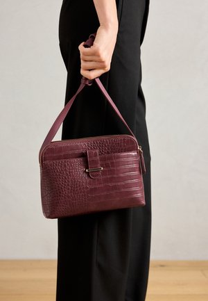 Hand houdt een kleine bordeauxrode krokodillenstructuur handtas vast met een rits en detail van de voorste riem tegen een zwarte broek en lichte achtergrond.