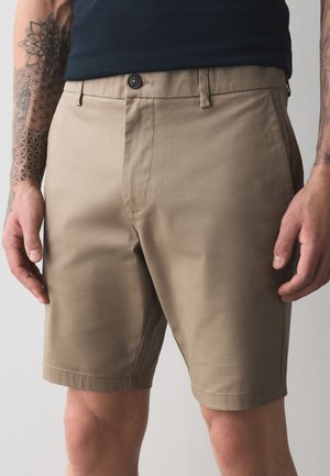 Mann trägt beige knielange Shorts und ein schwarzes Hemd, zeigt Tattoos auf beiden Unterarmen vor einfachem Hintergrund.