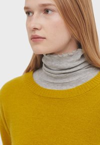 Maglione giallo lavorato a maglia con collo rotondo indossato sopra un dolcevita grigio chiaro con orlo ondulato. I capelli sono lisci e di lunghezza media.
