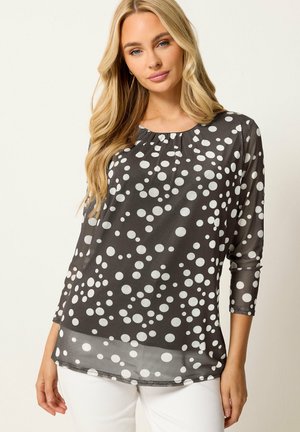Mujer con cabello largo y rubio que lleva una blusa negra con lunares blancos y mangas tres cuartos, combinada con pantalones blancos, de pie frente a un fondo liso.