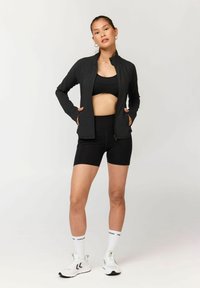 Donna con reggiseno sportivo nero, giacca nera, pantaloncini neri, calzini bianchi con scritta e scarpe da ginnastica bianche, in piedi davanti a uno sfondo chiaro uniforme.