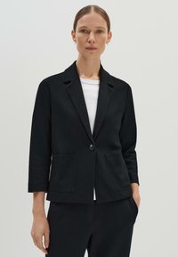 Zwarte blazer met een beneden de kraag, één knoopsluiting en twee voorzakken. Gepaard met een wit shirt onder de jas.