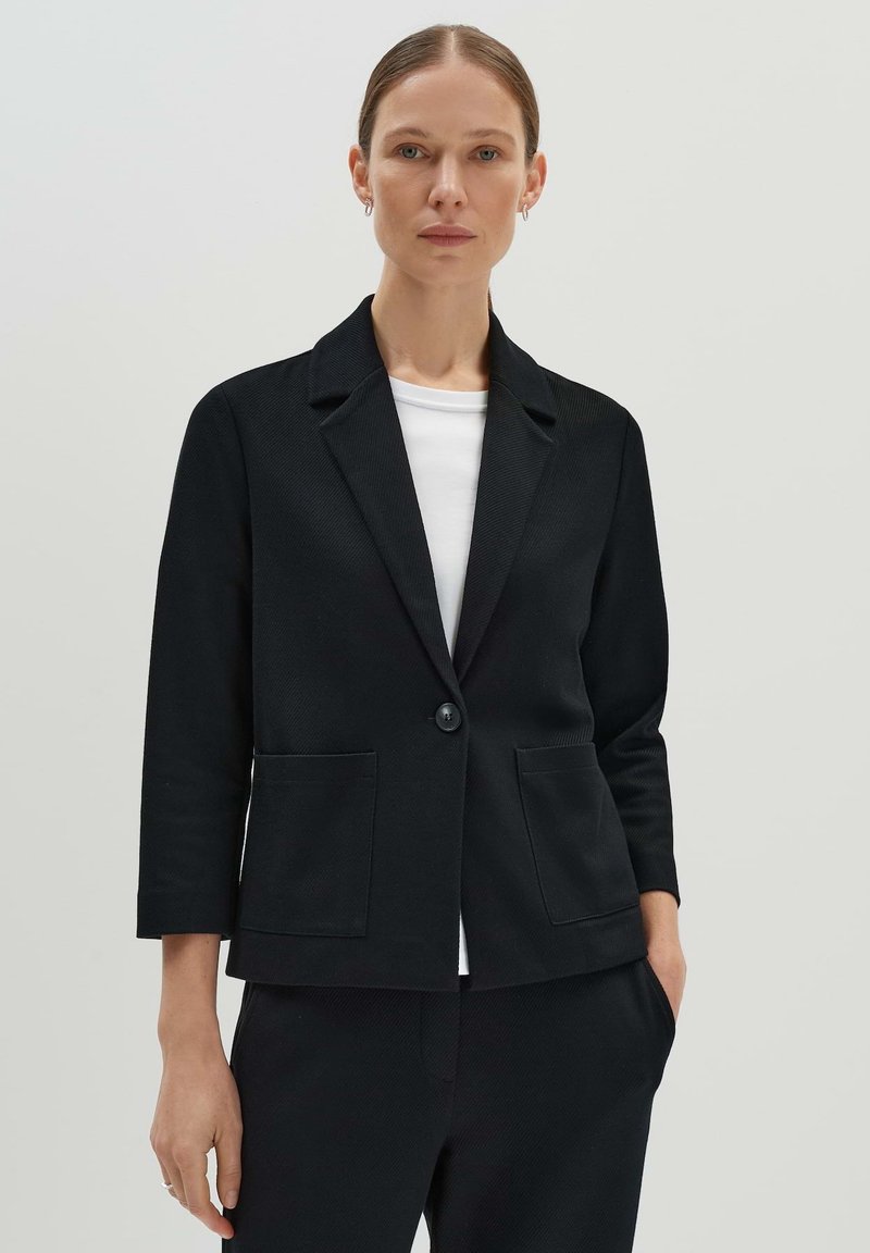 Zwarte blazer met een beneden de kraag, één knoopsluiting en twee voorzakken. Gepaard met een wit shirt onder de jas.