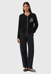 Donna che indossa un maglione nero con zip e logo bianco sul petto, jeans neri a gamba dritta e ballerine nere con fibbie argentate.