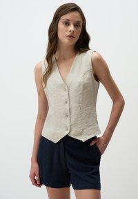 Gilet sans manches en lin beige clair avec un décollet�é en V, cinq boutons sur le devant et un ourlet légèrement arrondi. Associé à un short foncé.