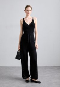 Top in maglia nera a costine con scollatura a V, abbinato a pantaloni neri larghi con vita alta e cintura annodabile. Gli accessori includono una borsa a tracolla nera intrecciata.