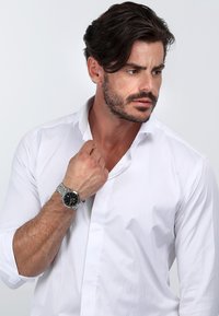 Hvid skjorte med knapkrave, båret med et sølvfarvet mesh armbåndsur med sort urskive og sølvfarvede detaljer.