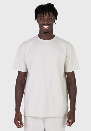 Uśmiechnięty młody mężczyzna z krótkimi dredami, ubrany w prostą, białą, oversize'ową koszulkę i białe spodnie na jasnoszarym tle.