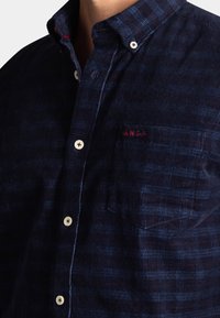 Navy corduroy shirt met een button-down kraag, met een borstzak en een rood geborduurd logo. Witte knopen contrasteren met de stof.