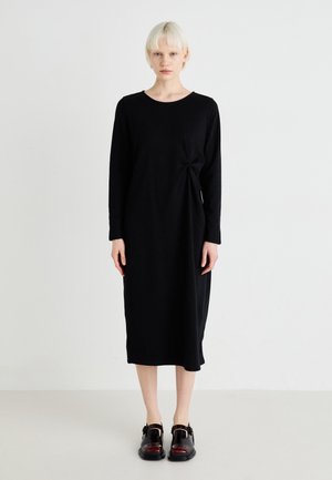 Vestido negro de manga larga con un corte relajado, que presenta un detalle de nudo en el lado y una textura suave, combinado con mocasines oscuros y gruesos.