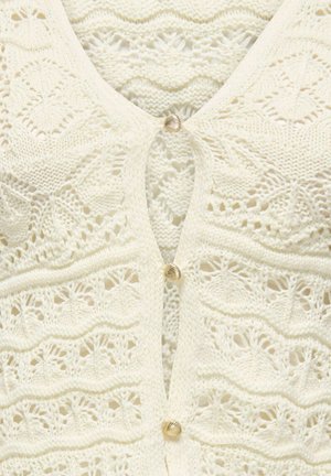 Cardigan tricoté couleur crème avec des motifs de dentelle complexes et de petits boutons dorés en forme de coquillage le long de l'avant.