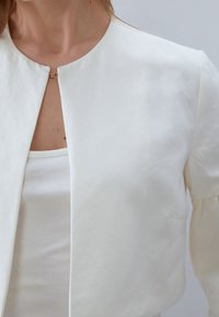 Chaqueta blanca recortada, textura suave, escote redondo, cierre de un solo broche dorado, sutil detalle de costura, superpuesta sobre un top blanco sin mangas.