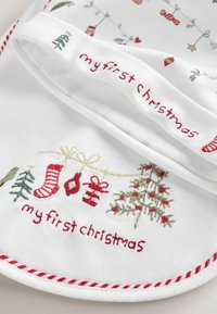 Hvid bomuldsforklæde med rød og grøn broderi, der står "min første jul", med festlige mønstre som træer, strømper og gaver.