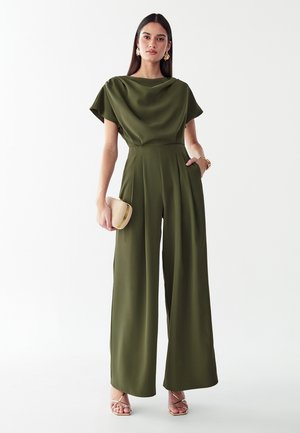 Femme debout portant une combinaison pantalon kaki à jambes larges, tenant une pochette beige, avec des boucles d’oreilles dorées et des talons ouverts sur fond blanc uni.