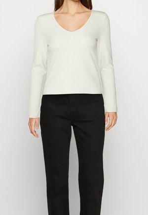 Kobieta ubrana w kremowy, długi sweter na guziki z dekoltem w serek i czarne, wysokie spodnie o prostych nogawkach, stojąca na białym tle.