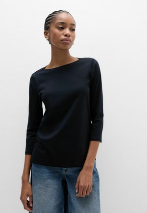 Frau mit geflochtenem Haar, die ein schwarzes Oberteil mit Dreiviertelärmeln und blaue Jeans trägt und vor einem schlichten weißen Hintergrund steht.