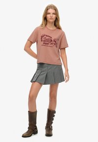 Joven mujer con una camiseta gráfica rosa polvorienta, falda plisada de rayas grises y botas altas marrones con hebilla, sobre un fondo blanco.