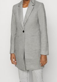 Abrigo gris a medida con un único botón negro, solapas con muesca, dos bolsillos delanteros y tela texturizada. Llevado sobre una camisa blanca.