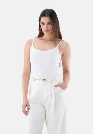 Donna con lunghi capelli castani che indossa un top bianco senza maniche e pantaloni bianchi a vita alta con cintura a catena dorata, in piedi con la mano in tasca.