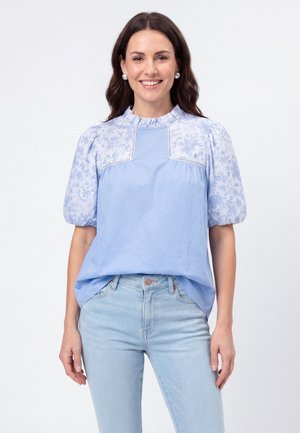 Frau lächelt, trägt hellblaue Bluse mit floralen Puffärmeln und Rüschenkragen, kombiniert mit hellblauen Jeans, steht vor weißem Hintergrund.