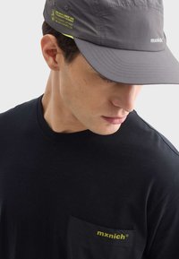 Šedá kšiltovka s kšiltem, s žlutými akcenty a brandingem. Černé tričko s malou kapsou označenou "mxnich." Jednoduchý, minimalistický design.