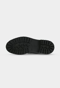 Semelle de chaussure en caoutchouc noir avec des rainures profondes et des motifs de grip en forme de croix sur un fond clair uni.