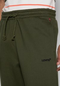 Olijfgroene sweatpants met een elastische tailleband en trekkoord, voorzien van een donker geborduurd logo op de dij. Glad, katoen materiaal.