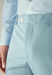 Lichtblauwe broeken van een soepele stof, met een sluiting aan de voorkant met een knoop en zijzakken, met strakke lijnen en subtiele stiksels.