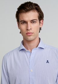 Hellblau gestreiftes Button-Down-Hemd aus Baumwollstoff, mit einem kleinen dunkelblauen Totenkopf- und Kreuzbones-Logo auf der linken Brustseite.
