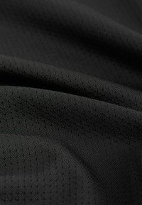 Tissu noir avec de petites perforations drapé en plis doux, montrant la texture et un matériau respirant semblable à un filet.
