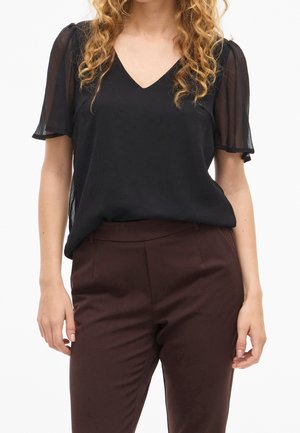 Femme portant un chemisier noir transparent à manches courtes avec un col en V et un pantalon ajusté marron foncé, debout avec les bras détendus le long du corps.