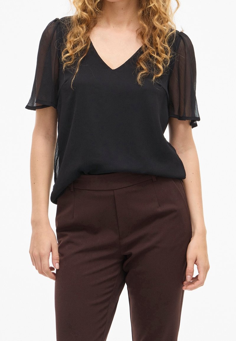 Femme portant un chemisier noir transparent à manches courtes avec un col en V et un pantalon ajusté marron foncé, debout avec les bras détendus le long du corps.