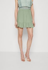 Jupe verte clair à volants avec une taille élastique, associée à un crop top texturé. Le mannequin porte des sandales à talons hauts nudes avec des lanières.