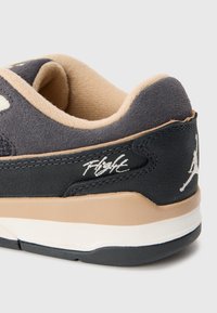 Primer plano del talón de una sneaker negra y beige con el texto "Flight" bordado y el logo Jumpman blanco sobre un fondo limpio.