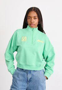 Felpa verde menta con zip e collo alto, grande numero ricamato 59 e dettagli con logo. Vestibilità comoda con orlo e polsini a costine.