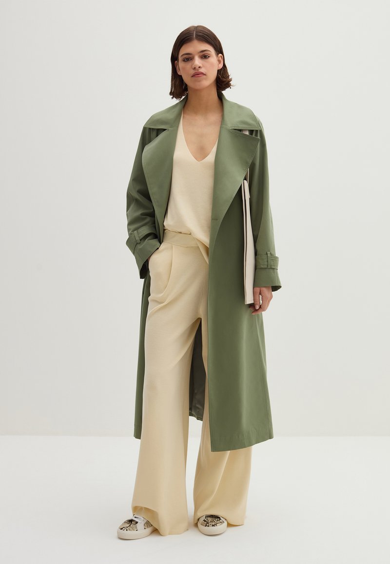 Donna in piedi che indossa un cappotto lungo verde oliva, un top beige a scollo a V, pantaloni beige a gamba larga e sneakers con stampa pitone, mentre tiene una clutch beige.