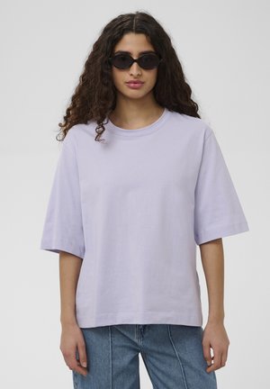 Kvinde med krøllet hår iført sorte solbriller, en løs lys lilla t-shirt med korte ærmer og blå jeans, stående mod en ensfarvet baggrund.