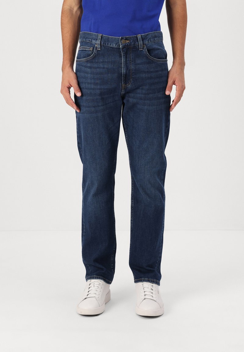 Dunkelblaue Jeans aus Denim mit geradem Bein, Fünf-Taschen-Design und subtilen Abnutzungserscheinungen auf der Vorderseite. Getragen mit weißen Sneakers.