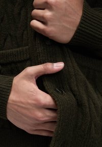 Dunkelolivgrüner Strickcardigan mit einem strukturierten Muster, gerippten Bündchen und schwarzen Knöpfen, während Hände den Stoff festhalten.