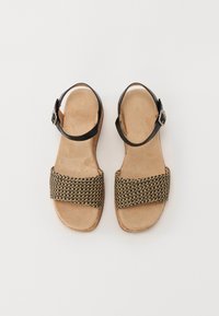 Svarte skinnsandaler med et vevd brunt stropp, flat såle og ankelspenne, med en myk beige innersåle og teksturert overflate.