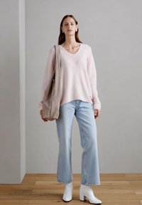 Lichtroze oversized trui in zachte stof, rechte blauwe jeans en witte enkellaarsjes. Beige tote bag over de schouder.