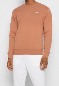 Brun sweatshirt i mjukt material med långa ärmar, rund halsringning och liten vit Nike-logga på bröstet. Matchad med vita byxor.