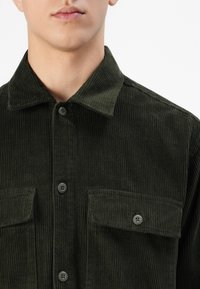 Homme portant une chemise en velours côtelé vert foncé avec poches à rabat, gros plan montrant le col et la zone de la poitrine.