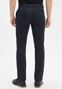 Mężczyzna ubrany w ciemnoniebieskie, slim-fit spodnie oraz czarne skórzane mokasyny, stoi plecami do widza na tle jednolitego białego tła.